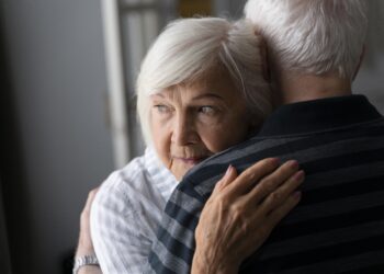 Older woman with short white hair hugs an older man, resting her head on his shoulder, looking sideways.