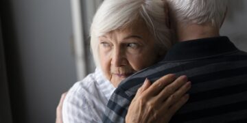 Older woman with short white hair hugs an older man, resting her head on his shoulder, looking sideways.