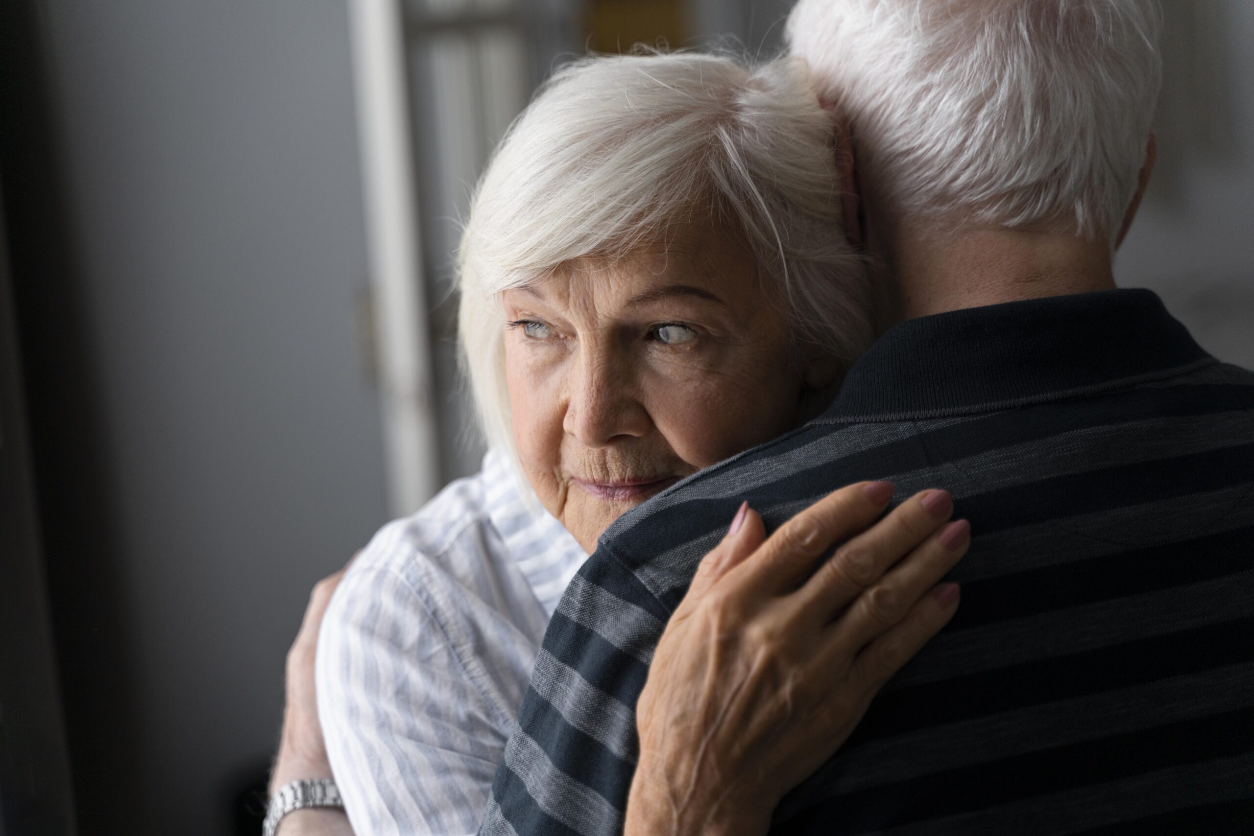 Older woman with short white hair hugs an older man resting her head on his shoulder looking sideways