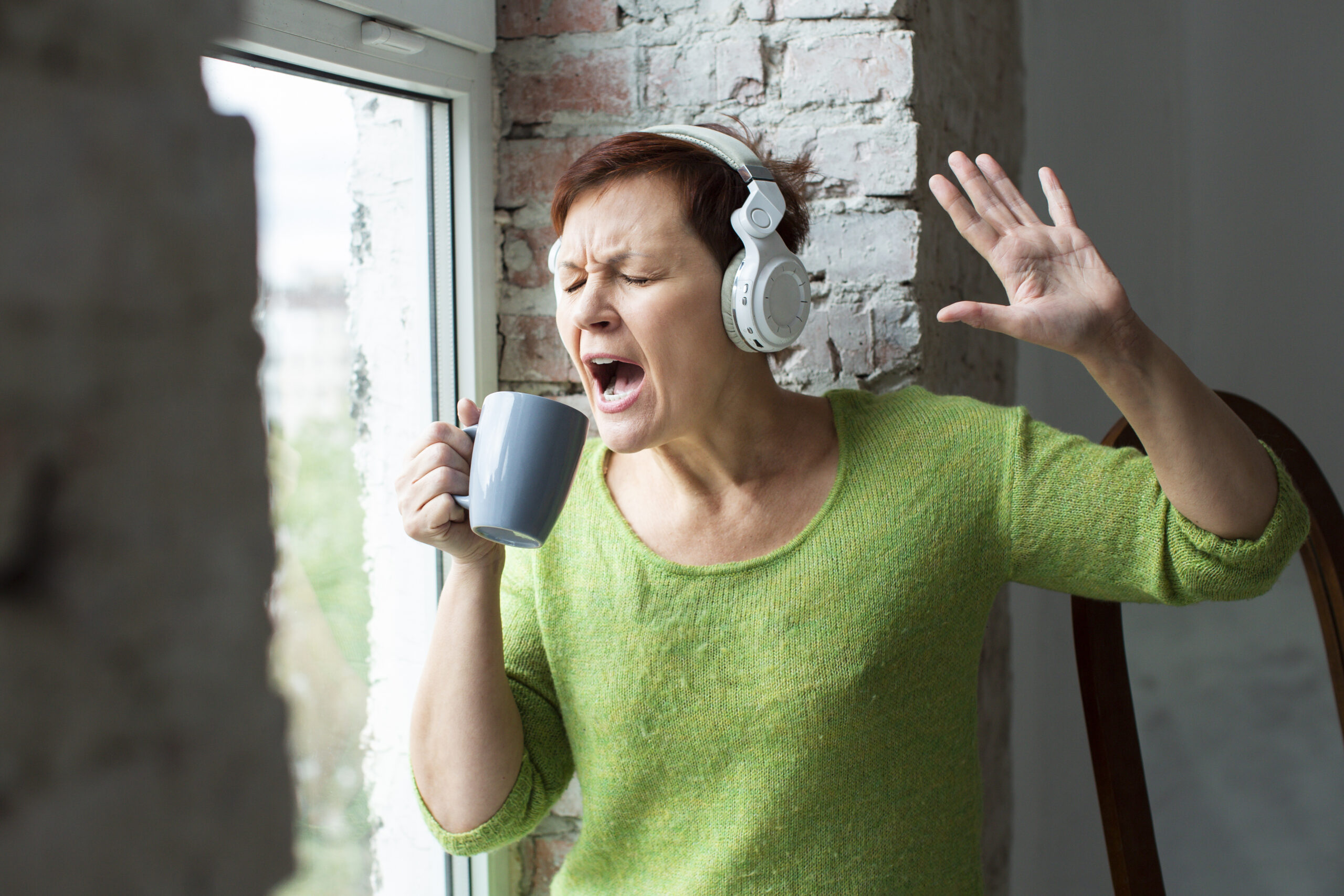 Woman with white over ear headphones sings into a mug near a window wearing a bright green sweater
