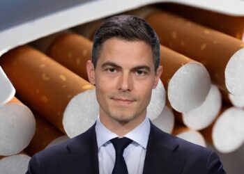 Portrait of a man in a dark suit and tie set against a backdrop of cigarettes stacked behind him