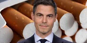 Portrait of a man in a dark suit and tie, set against a backdrop of cigarettes stacked behind him.