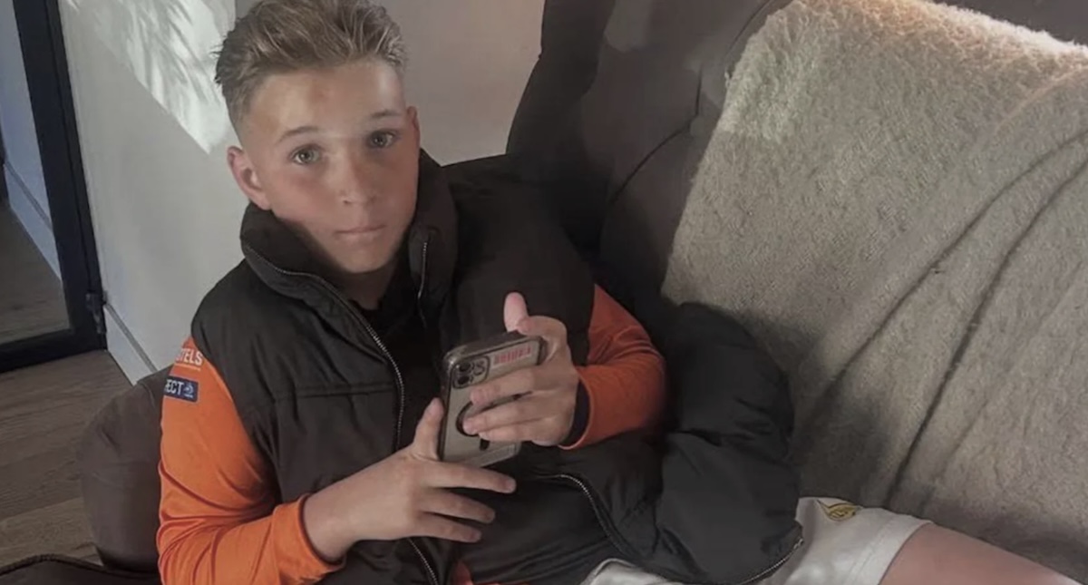 Young person sitting on a sofa wearing an orange long sleeve shirt and black vest looking at a smartphone in their hands