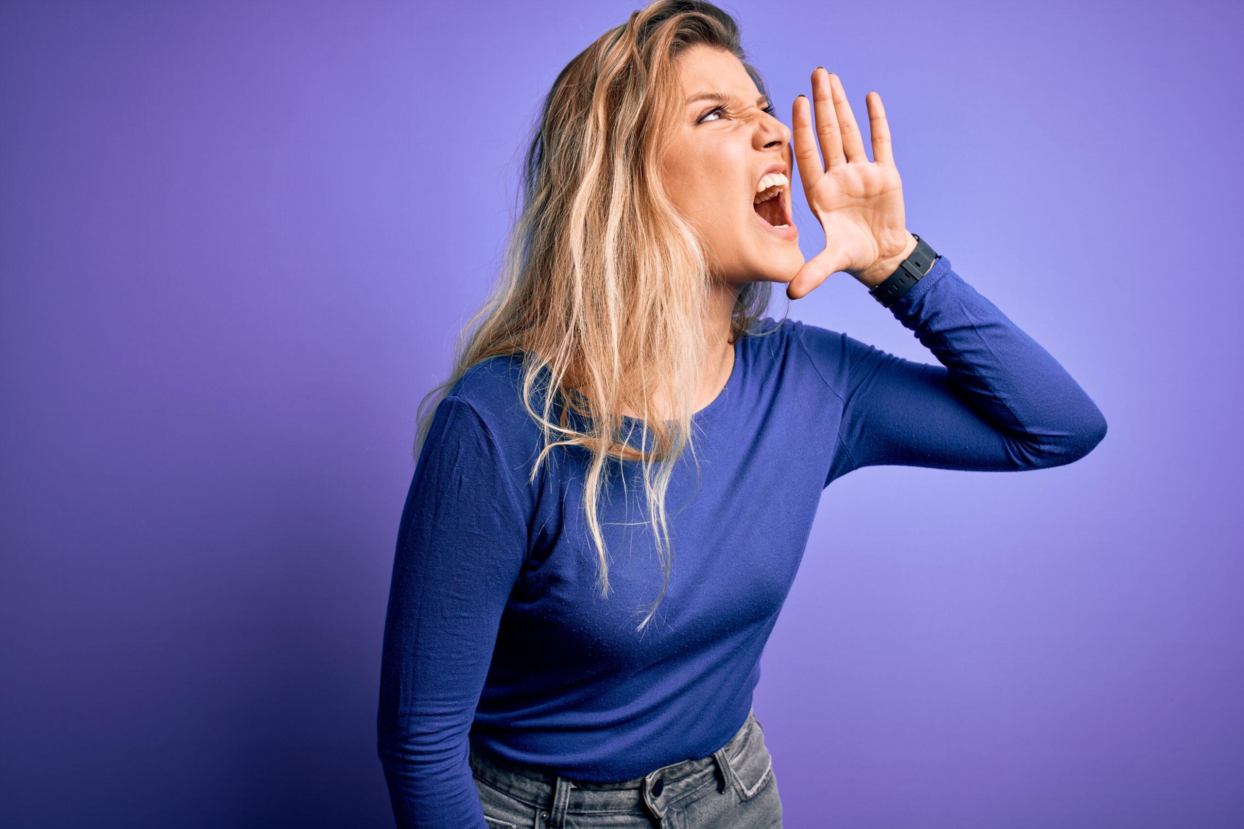 Woman with long blond hair in a blue shirt shouting with her hand cupped near her mouth against a purple background