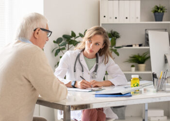 Female doctor with stethoscope records notes for an older male patient at a bright clinic desk computer nearby