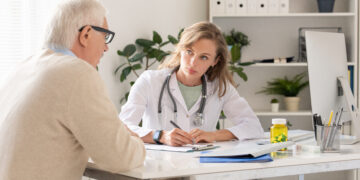 Female doctor with stethoscope records notes for an older male patient at a bright clinic desk, computer nearby.
