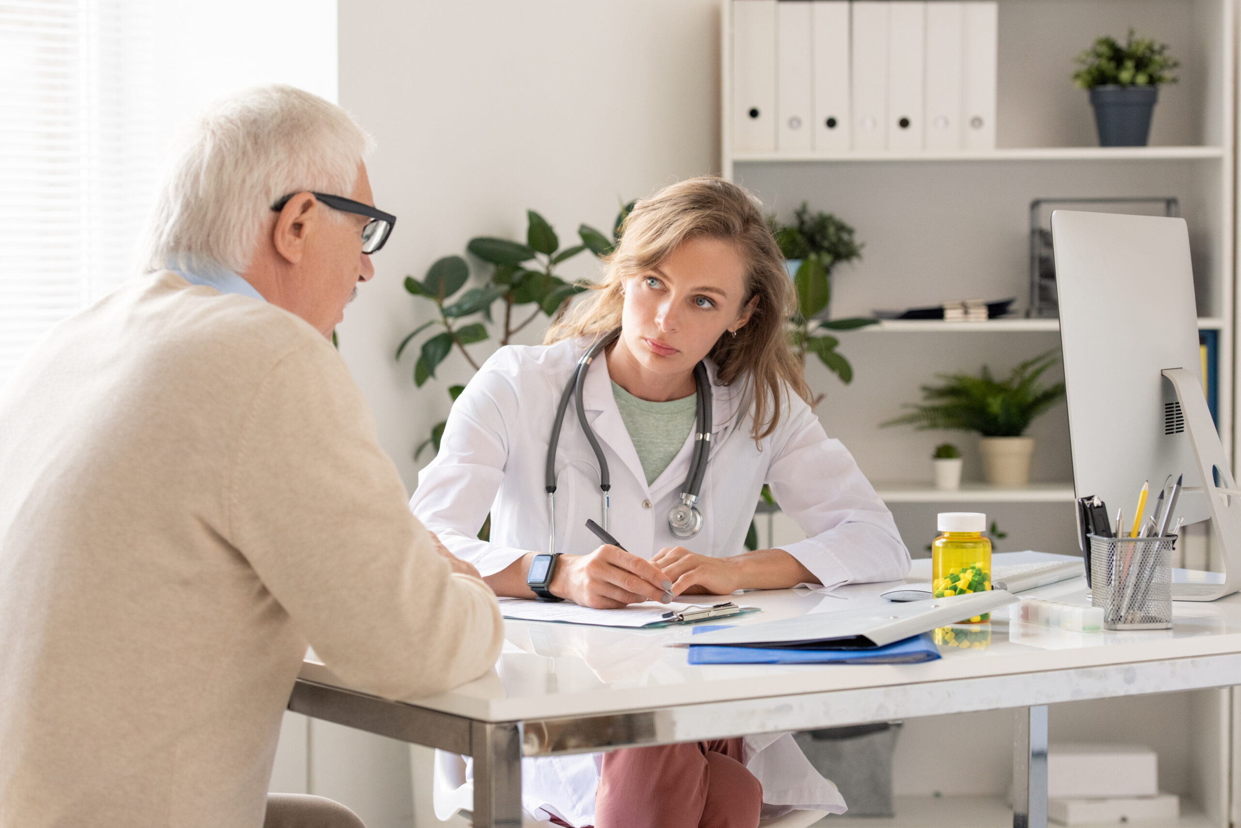 Female doctor with stethoscope records notes for an older male patient at a bright clinic desk computer nearby