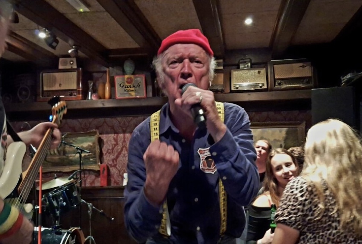Male singer in a red beret performs into a mic at a cozy bar a tape measure draped around his neck while musicians and patrons watch