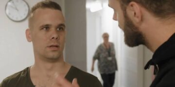 Two men in a hallway talking, with a clock on the wall and a blurred person walking in the background.
