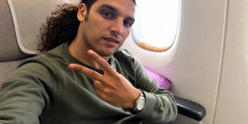 Man with long curly hair wearing a green sweatshirt sits in an airplane seat near a window, making a peace sign with his fingers.