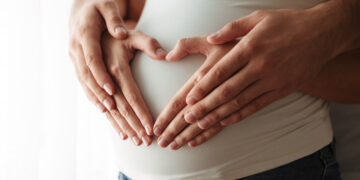 Close-up of a pregnant belly being cradled by two hands forming a heart shape over the abdomen