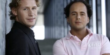 Two men pose in an industrial hallway; one in a black blazer leans against a wall, the other in a pink polo with arms crossed.
