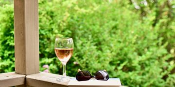 Wine glass of rosé, black sunglasses, and a closed book resting on a wooden railing outdoors, with a lush green garden in the background.