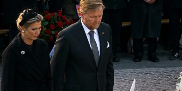 Two adults in formal black attire standing near a red rose wreath at a funeral or memorial ceremony.