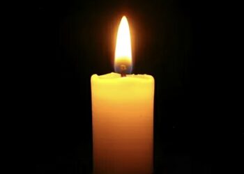 Tall yellow candle burning with a bright flame against a dark background, casting a warm glow.