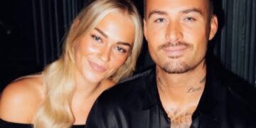 Smiling couple posing closely in a dark background; woman with long blonde hair in an off-the-shoulder black top, man with tattoos in a black shirt.