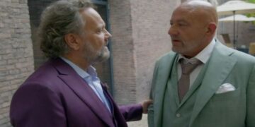 Two middle-aged men in colorful suits talking outdoors by a brick wall, one resting a hand on the other's arm.