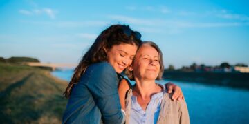 Two women share a warm hug by a riverside during golden hour, smiling at the camera (blue sky).