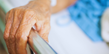 Elderly hand gripping a hospital bed rail with an IV in the forearm.
