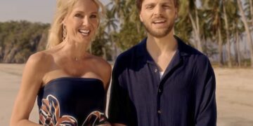 Smiling couple stands on a sunny beach with palm trees in the background, posing for the camera.
