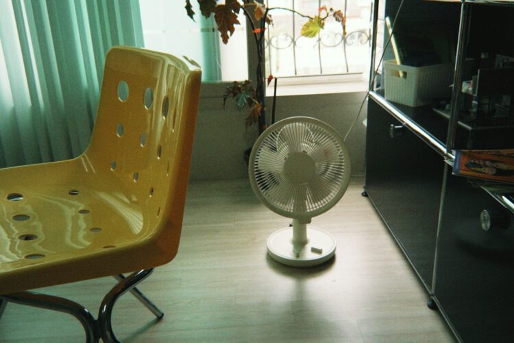 Yellow chair, white fan, and window in a room.