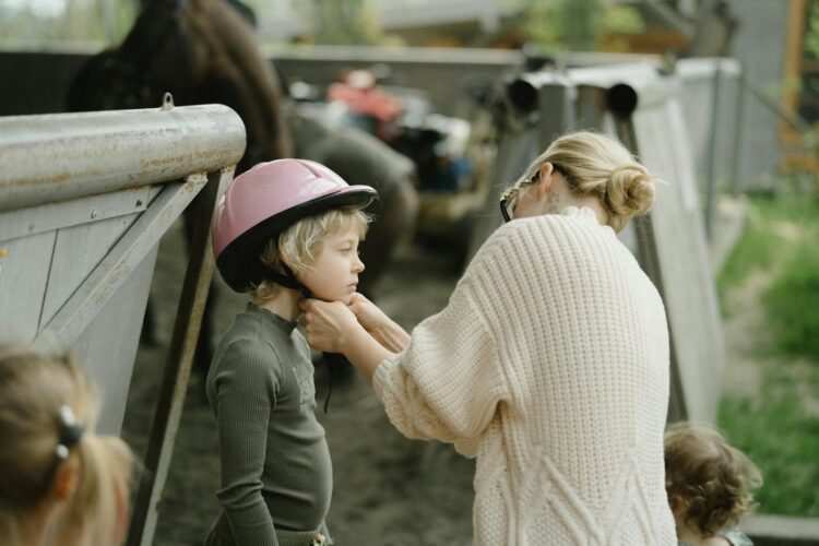 Mother helping child with a pink helmet in a farm setting, emphasizing care and safety.