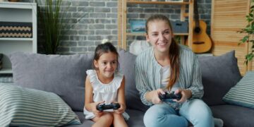 Mother and daughter playing video games together on sofa.